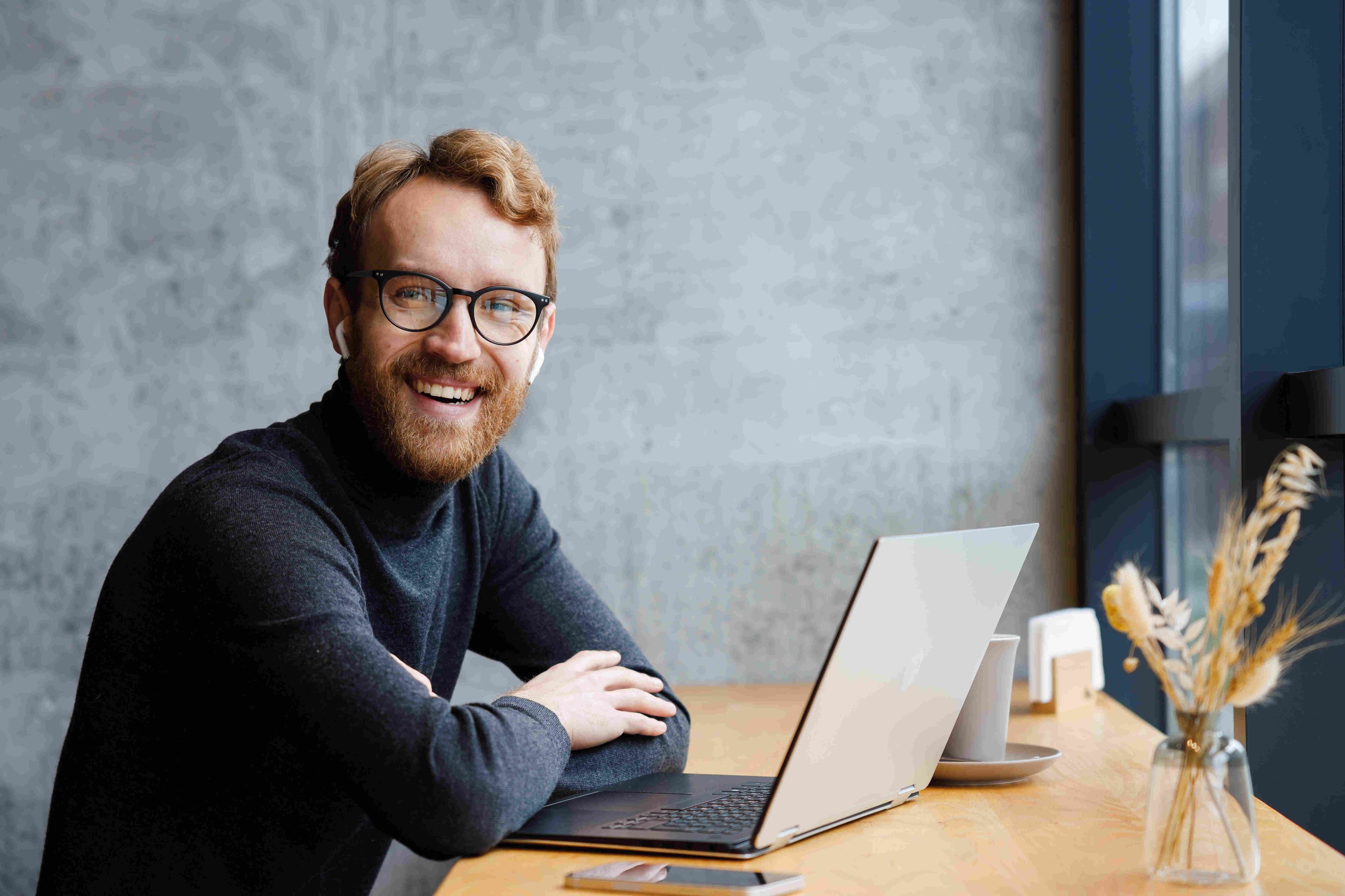 Young entrepreneur wearing glasses, working on a laptop in a stylish café. Smiling at the camera, he embodies the spirit of freelancing, remote work, and modern online communication for small businesses.
