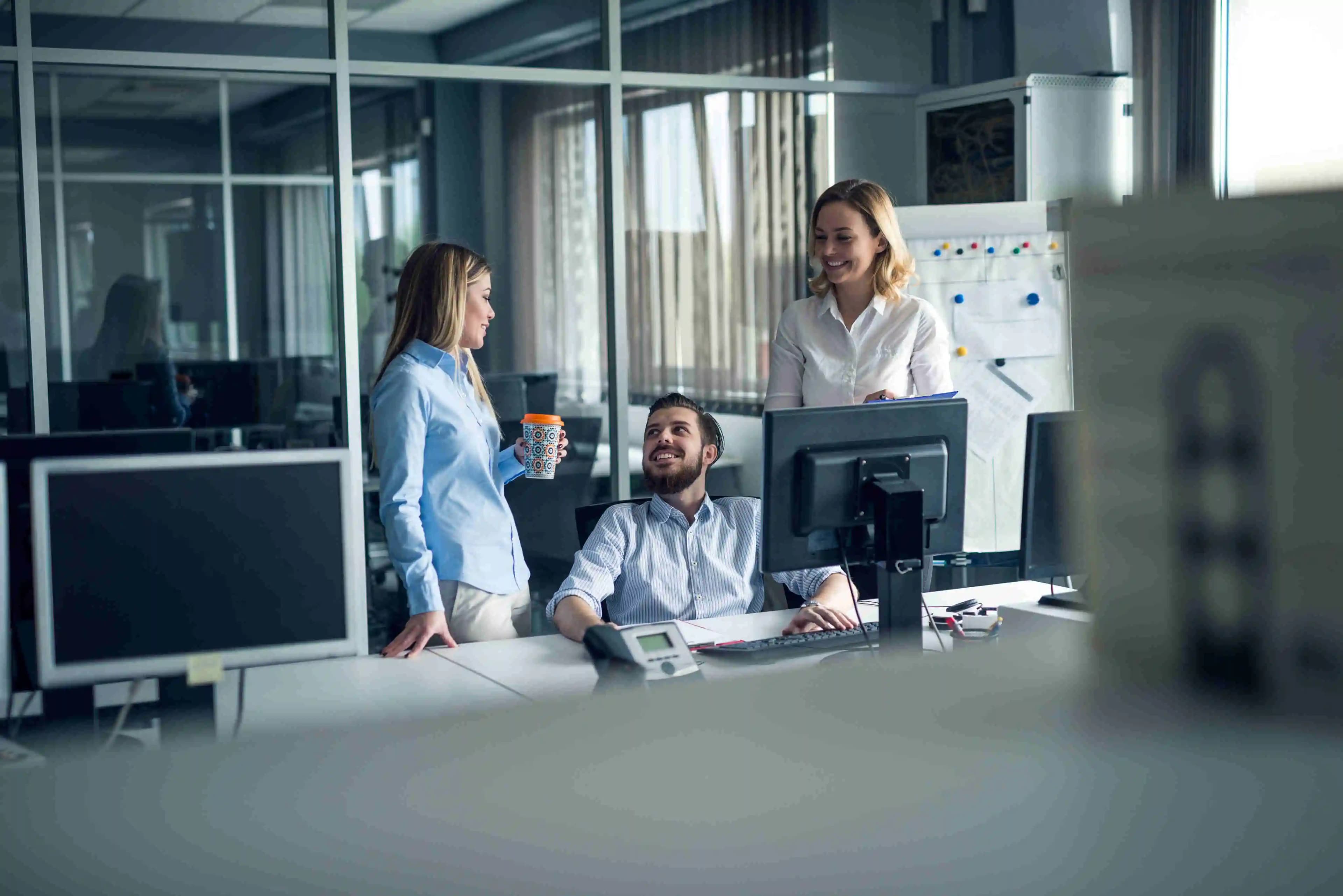 Three colleagues laughing and discussing work-related topics. One of them is holding a colorful thermal mug with coffee.
