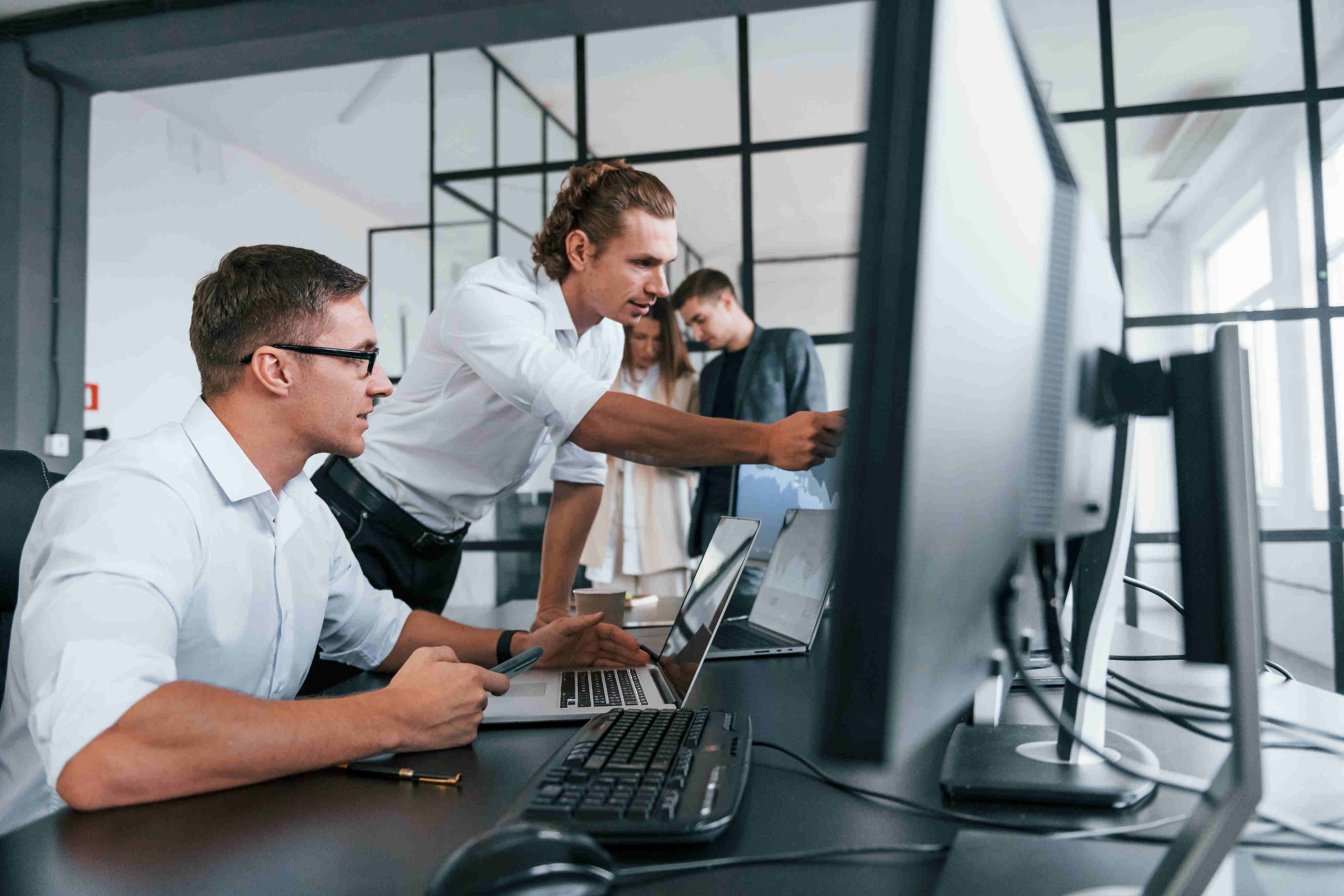Developers are collaboratively solving a task at a computer in a modern office. One of them is pointing out an important detail to a colleague.